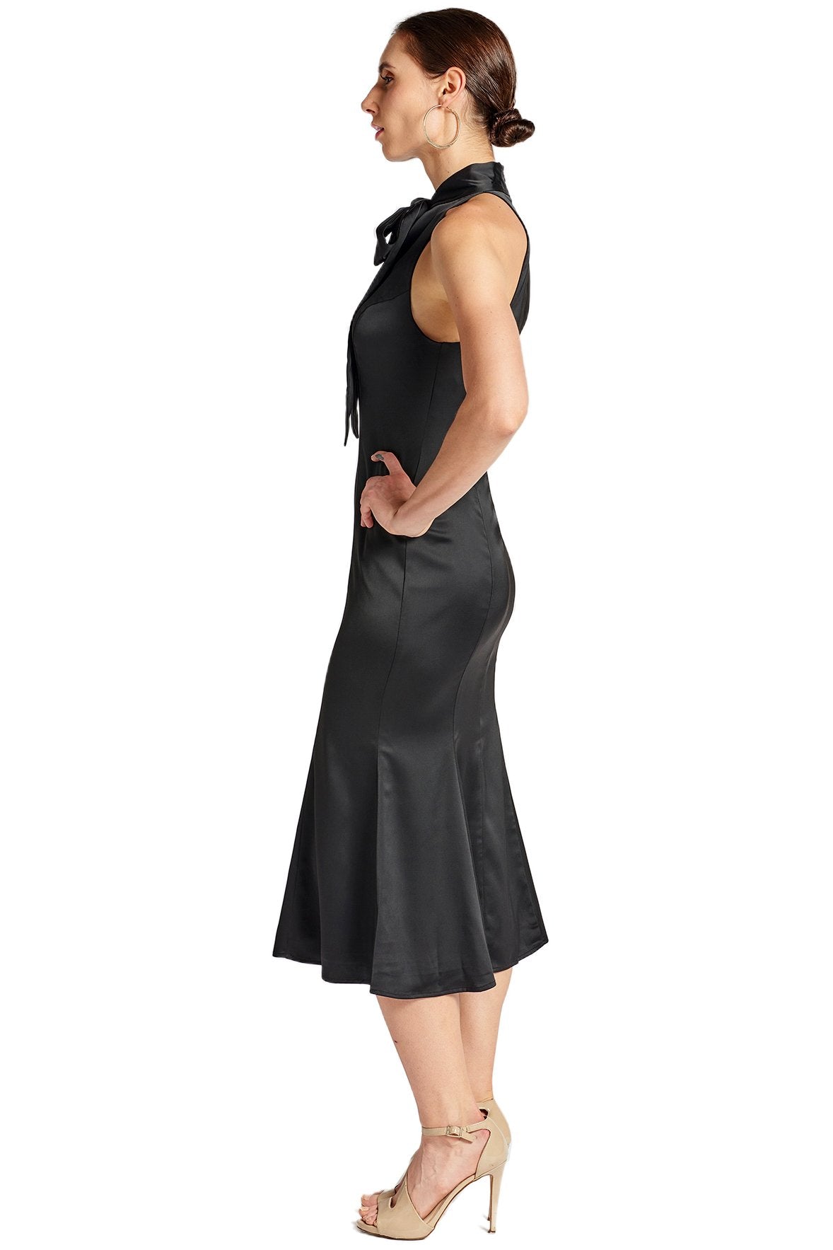Side view of model wearing black stretch sleeveless satin midi dress with pussy bow necktie and mermaid hem.