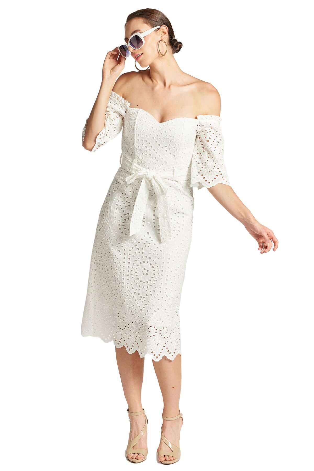 Front view of model wearing the Simona Maghen Jasmine Dress, white cotton scalloped eyelet midi a-line dress with off the shoulder sweetheart neckline, self belt, and short sleeves. Accessorized with a pair of big gold hoop earrings and white sunglasses.