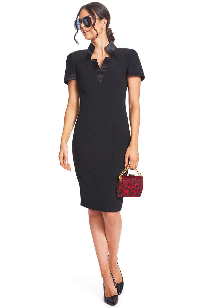 Front view of model wearing the Simona Maghen Top Notch Dress, notch neck high collar poly/spandex crepe sheath dress with split cap sleeves and satin contrast bands. Accessorized with a pair of black aviators and a red and black clutch with a gold chain.